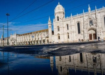 Un grande edificio storico in pietra bianca con un'architettura gotica decorata si riflette in una pozzanghera sulla strada. Il cielo è limpido e azzurro. Alcune persone stanno camminando vicino all'edificio mentre le linee del tram elettrico passano davanti ad esso, ricordando una giornata serena a Lisbona prima dell'allarme Tsunami.