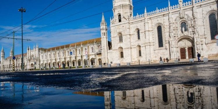Un grande edificio storico in pietra bianca con un'architettura gotica decorata si riflette in una pozzanghera sulla strada. Il cielo è limpido e azzurro. Alcune persone stanno camminando vicino all'edificio mentre le linee del tram elettrico passano davanti ad esso, ricordando una giornata serena a Lisbona prima dell'allarme Tsunami.