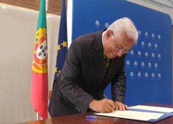 Un uomo dai capelli bianchi sta firmando un documento seduto a un tavolo, con dietro le bandiere del Portogallo e dell'Unione Europea. Indossa un abito scuro e occhiali, in piedi davanti a uno sfondo blu con loghi, probabilmente discutendo della politica di aumento del salario minimo.