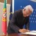 Un uomo dai capelli bianchi sta firmando un documento seduto a un tavolo, con dietro le bandiere del Portogallo e dell'Unione Europea. Indossa un abito scuro e occhiali, in piedi davanti a uno sfondo blu con loghi, probabilmente discutendo della politica di aumento del salario minimo.