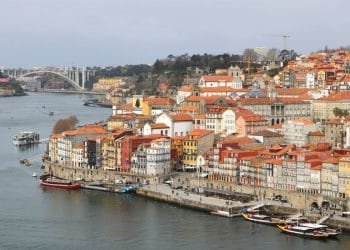 Veduta aerea di Porto, Portogallo, dove dove comprano casa stranieri portogallo mette in mostra gli edifici colorati della città con tetti di tegole rosse lungo il fiume Douro. Le barche sono attraccate lungo il fiume e sullo sfondo un grande ponte ad arco attraversa l'acqua. Il cielo è coperto.