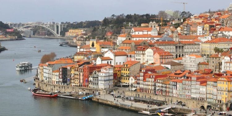 Veduta aerea di Porto, Portogallo, dove dove comprano casa stranieri portogallo mette in mostra gli edifici colorati della città con tetti di tegole rosse lungo il fiume Douro. Le barche sono attraccate lungo il fiume e sullo sfondo un grande ponte ad arco attraversa l'acqua. Il cielo è coperto.