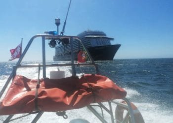 Una grande nave da crociera naviga sull'oceano sotto un cielo azzurro, vista dal ponte posteriore di una barca più piccola. L'imbarcazione più piccola, che potrebbe appartenere alla marina militare portoghese, è dotata di dotazioni di sicurezza tra cui un salvagente arancione e bandiere. Il mare è calmo con onde dolci.