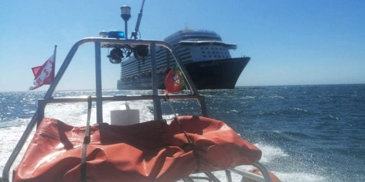 Una grande nave da crociera naviga sull'oceano sotto un cielo azzurro, vista dal ponte posteriore di una barca più piccola. L'imbarcazione più piccola, che potrebbe appartenere alla marina militare portoghese, è dotata di dotazioni di sicurezza tra cui un salvagente arancione e bandiere. Il mare è calmo con onde dolci.