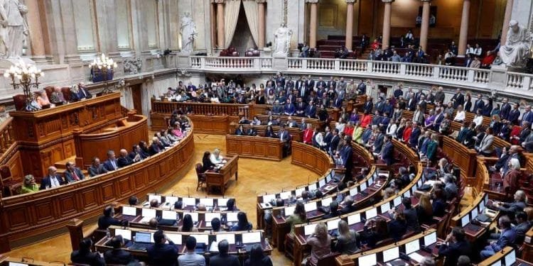 Una sessione parlamentare all'interno di un'aula magna ricca di soffitti alti e statue. I membri del parlamento, che discutono della modifica tasse pensionati Portogallo, sono seduti in file curve davanti a scrivanie con schermi, mentre un oratore si rivolge loro da una posizione elevata. Anche la tribuna pubblica in alto è occupata.