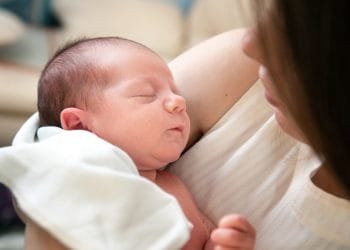 Un primo piano di un neonato addormentato cullato tra le braccia di un adulto. Il bambino, uno dei nomi bambini Portogallo, ha la pelle chiara e i capelli corti. L'adulto, il cui volto è parzialmente visibile, indossa una camicia bianca, tiene delicatamente stretto il bambino con un morbido panno bianco sotto la testa del bambino.