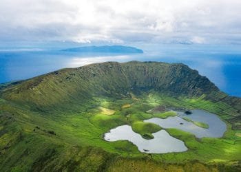 Veduta aerea del cratere vulcanico dell'isola di Corvo nelle Azzorre, Portogallo. Il vasto cratere verde è punteggiato da piccoli laghi e circondato dal blu dell'Oceano Atlantico, con un'altra isola visibile in lontananza sotto un cielo parzialmente nuvoloso: una straordinaria novità voli Azzorre.