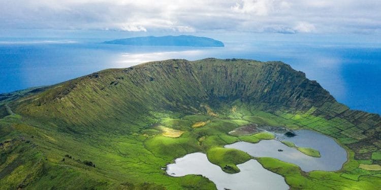 Veduta aerea del cratere vulcanico dell'isola di Corvo nelle Azzorre, Portogallo. Il vasto cratere verde è punteggiato da piccoli laghi e circondato dal blu dell'Oceano Atlantico, con un'altra isola visibile in lontananza sotto un cielo parzialmente nuvoloso: una straordinaria novità voli Azzorre.