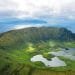 Veduta aerea del cratere vulcanico dell'isola di Corvo nelle Azzorre, Portogallo. Il vasto cratere verde è punteggiato da piccoli laghi e circondato dal blu dell'Oceano Atlantico, con un'altra isola visibile in lontananza sotto un cielo parzialmente nuvoloso: una straordinaria novità voli Azzorre.
