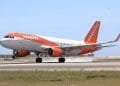 Un aereo EasyJet, dipinto nei colori bianco e arancione caratteristici della compagnia aerea, sta decollando o atterrando su una pista con una leggera scia di fumo visibile dai pneumatici. Il cielo limpido e le strutture aeroportuali sullo sfondo suggeriscono un'altra giornata impegnativa per i voli EasyJet in Portogallo.