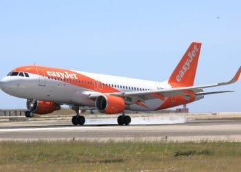 Un aereo EasyJet, dipinto nei colori bianco e arancione caratteristici della compagnia aerea, sta decollando o atterrando su una pista con una leggera scia di fumo visibile dai pneumatici. Il cielo limpido e le strutture aeroportuali sullo sfondo suggeriscono un'altra giornata impegnativa per i voli EasyJet in Portogallo.