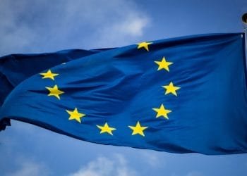 L'immagine mostra la bandiera dell'Unione Europea, che è blu con un cerchio di dodici stelle gialle al centro. La bandiera sventola al vento sullo sfondo di un cielo parzialmente nuvoloso, a simboleggiare l'unità e la stabilità in mezzo alle discussioni sui tassi Euribor e alle politiche mutui del Portogallo.