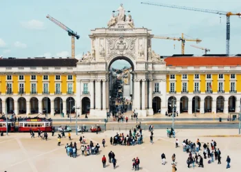 Lisbona, praca do comercio è cuore del turismo in Portogallo ospita l'Arco da Rua Augusta, un grande arco architettonico con sculture intricate. Edifici gialli con finestre ad arco fiancheggiano l'arco, mentre le gru incombono sullo sfondo. I tram e i gruppi di persone contribuiscono alla scena vivace.