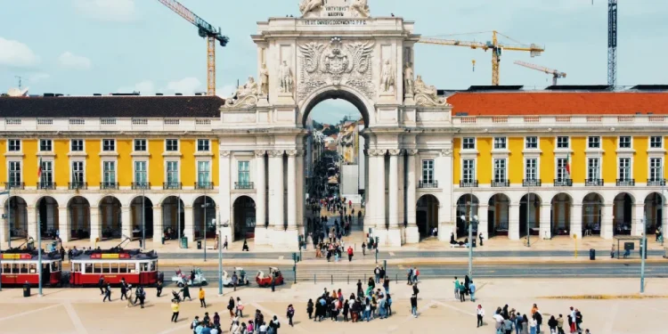 Lisbona, praca do comercio è cuore del turismo in Portogallo ospita l'Arco da Rua Augusta, un grande arco architettonico con sculture intricate. Edifici gialli con finestre ad arco fiancheggiano l'arco, mentre le gru incombono sullo sfondo. I tram e i gruppi di persone contribuiscono alla scena vivace.