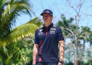 Un uomo con una maglietta e un berretto della squadra di corse blu navy si trova all'aperto sotto un cielo azzurro brillante con palme sullo sfondo, possibilmente in una località tropicale. Sembra che stia discutendo di un incidente di autonoleggio negato o condividendo storie sull'ultima gara di Verstappen.