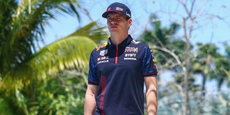 Un uomo con una maglietta e un berretto della squadra di corse blu navy si trova all'aperto sotto un cielo azzurro brillante con palme sullo sfondo, possibilmente in una località tropicale. Sembra che stia discutendo di un incidente di autonoleggio negato o condividendo storie sull'ultima gara di Verstappen.