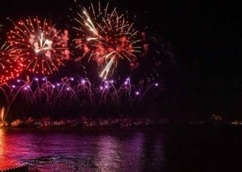 Un vibrante spettacolo pirotecnico illumina il cielo notturno di Lisbona con esplosioni di tonalità rosse, viola e arancioni che si riflettono sull'acqua scura sottostante. I fuochi d'artificio creano una serie di motivi e colori sullo sfondo nero, catturando lo spirito festoso del Capodanno Portogallo.