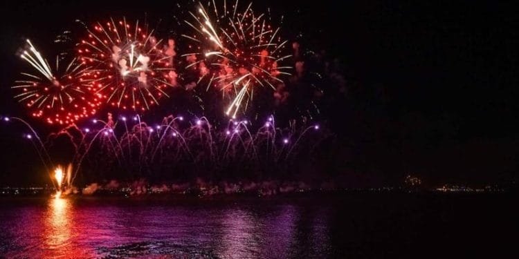 Un vibrante spettacolo pirotecnico illumina il cielo notturno di Lisbona con esplosioni di tonalità rosse, viola e arancioni che si riflettono sull'acqua scura sottostante. I fuochi d'artificio creano una serie di motivi e colori sullo sfondo nero, catturando lo spirito festoso del Capodanno Portogallo.