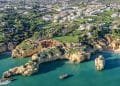 Veduta aerea della costa dell'Algarve, in Portogallo, caratterizzata da scogliere frastagliate, acque oceaniche color acquamarina e una spiaggia tranquilla. Il paesaggio sopra le scogliere è costellato di verde e un mix di edifici bianchi, mostrando una pittoresca miscela di bellezze naturali e aree residenziali, catturando davvero l'essenza del costo case dell'Algarve.