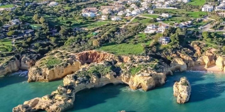 Veduta aerea della costa dell'Algarve, in Portogallo, caratterizzata da scogliere frastagliate, acque oceaniche color acquamarina e una spiaggia tranquilla. Il paesaggio sopra le scogliere è costellato di verde e un mix di edifici bianchi, mostrando una pittoresca miscela di bellezze naturali e aree residenziali, catturando davvero l'essenza del costo case dell'Algarve.