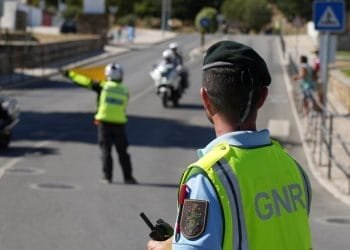 Un agente di polizia con berretto verde e gilet giallo con la scritta "GNR" dirige il traffico su una strada, rispettando la legge moto portogallo. Un altro agente con un gilet giallo segnala ai veicoli in arrivo, mentre due agenti in motocicletta vengono fermati nelle vicinanze. I pedoni stanno sul marciapiede osservando la scena.