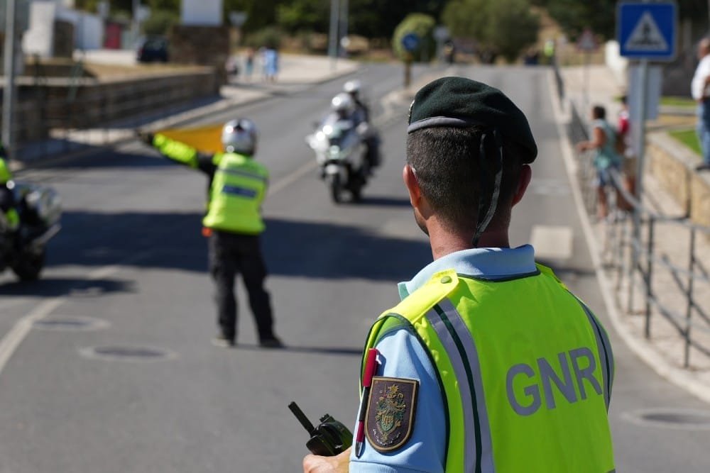 Un agente di polizia con berretto verde e gilet giallo con la scritta "GNR" dirige il traffico su una strada, rispettando la legge moto portogallo. Un altro agente con un gilet giallo segnala ai veicoli in arrivo, mentre due agenti in motocicletta vengono fermati nelle vicinanze. I pedoni stanno sul marciapiede osservando la scena.