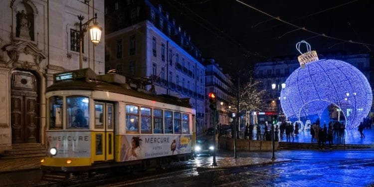 Un tram giallo attraversa una strada bagnata e riflettente di notte in una città. Lo sfondo presenta un grande ornamento natalizio blu illuminato ed edifici adornati con luci festive. Le persone si riuniscono attorno all'ornamento, godendosi l'atmosfera vacanziera, abbracciando le tradizioni natalizie del Portogallo.