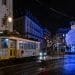 Un tram giallo attraversa una strada bagnata e riflettente di notte in una città. Lo sfondo presenta un grande ornamento natalizio blu illuminato ed edifici adornati con luci festive. Le persone si riuniscono attorno all'ornamento, godendosi l'atmosfera vacanziera, abbracciando le tradizioni natalizie del Portogallo.