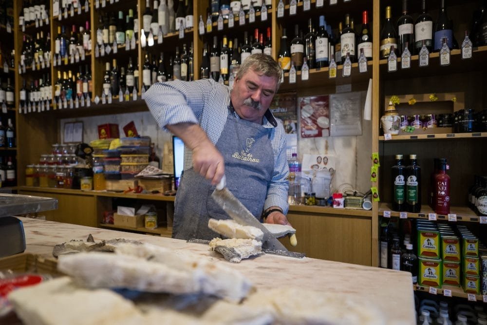 Un negoziante in camicia a quadri e grembiule affetta un grosso blocco di pesce essiccato e salato davanti al bancone di legno di un negozio pieno di bottiglie di vino e vari prodotti alimentari sugli scaffali dietro di lui. Il negozio caldo e disordinato evoca le tradizioni natalizie del Portogallo, facendolo sentire allo stesso tempo accogliente e nostalgico.