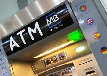 Primo piano di un bancomat. Il pannello superiore visualizza "ATM" e "MB MULTIBANCO". A destra ci sono le opzioni della lingua indicate dalle bandiere: portoghese, inglese, spagnolo, tedesco e francese. Lo schermo è vuoto e la macchina è dotata di pulsanti standard e slot per carte per operazioni come versare contanti attraverso bancomat.
