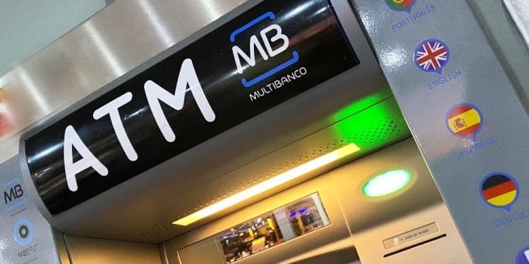 Primo piano di un bancomat. Il pannello superiore visualizza "ATM" e "MB MULTIBANCO". A destra ci sono le opzioni della lingua indicate dalle bandiere: portoghese, inglese, spagnolo, tedesco e francese. Lo schermo è vuoto e la macchina è dotata di pulsanti standard e slot per carte per operazioni come versare contanti attraverso bancomat.