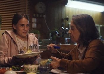 Una giovane donna con lunghi capelli scuri, che indossa una felpa con cappuccio rosa, tiene in mano un piatto mentre una donna anziana con un cardigan marrone le serve il cibo con una grande forchetta. Sono seduti a un tavolo da pranzo in una cucina accogliente e scarsamente illuminata, che ricorda le scene di un video di campagna sociale di grande impatto. Sul tavolo ci sono varie ciotole e piatti.