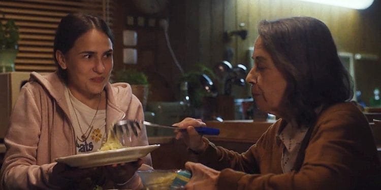 Una giovane donna con lunghi capelli scuri, che indossa una felpa con cappuccio rosa, tiene in mano un piatto mentre una donna anziana con un cardigan marrone le serve il cibo con una grande forchetta. Sono seduti a un tavolo da pranzo in una cucina accogliente e scarsamente illuminata, che ricorda le scene di un video di campagna sociale di grande impatto. Sul tavolo ci sono varie ciotole e piatti.