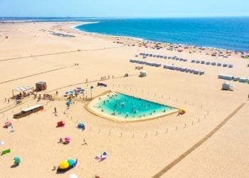 Veduta aerea di una spiaggia sabbiosa con una piccola *piscina acqua salata* recintata, piena di gente al centro. Ombrelli colorati punteggiano la zona e l'oceano blu si estende fino all'orizzonte. In lontananza si possono vedere file di cabine sulla spiaggia e visitatori sparsi. Giornata luminosa e soleggiata.