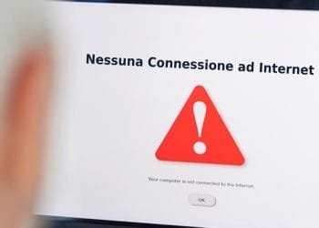 Lo schermo di un computer mostra un messaggio di errore in italiano che recita "Nessuna Connessione ad Internet" (Nessuna connessione Internet) con un triangolo rosso contenente un punto esclamativo al centro. Una persona sfocata alle prese con problemi di connessione casa sta visualizzando lo schermo.