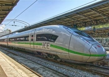 Un elegante treno ad alta velocità ICE (InterCity Express) color argento con una striscia verde è parcheggiato su un binario della stazione ferroviaria sotto una struttura coperta, che ricorda la realizzazione TGV portogallo. La scena è ben illuminata con il cielo sereno sullo sfondo.