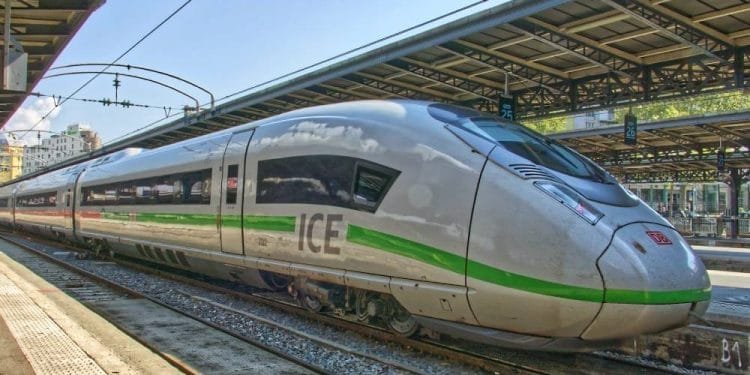 Un elegante treno ad alta velocità ICE (InterCity Express) color argento con una striscia verde è parcheggiato su un binario della stazione ferroviaria sotto una struttura coperta, che ricorda la realizzazione TGV portogallo. La scena è ben illuminata con il cielo sereno sullo sfondo.