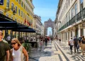 Una scena di strada a Lisbona con persone che camminano lungo rua Augusta, l'essenza del turismo in Portogallo. Si vedono edifici colorati con caffè all'aperto costeggiano la strada, conducendo all'Arco da rua Augusta in lontananza sotto un cielo azzurro e limpido.