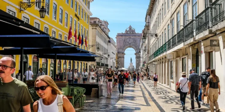 Una scena di strada a Lisbona con persone che camminano lungo rua Augusta, l'essenza del turismo in Portogallo. Si vedono edifici colorati con caffè all'aperto costeggiano la strada, conducendo all'Arco da rua Augusta in lontananza sotto un cielo azzurro e limpido.