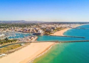 Veduta aerea di una città costiera dell'Algarve, portogallo, caratterizzata da un grande porto turistico con numerose barche ormeggiate, spiagge sabbiose che si estendono lungo la costa e un mix di edifici sparsi in tutta la città. L'oceano è di un turchese vibrante e il cielo è limpido, ideale per avventure in multiproprietà.