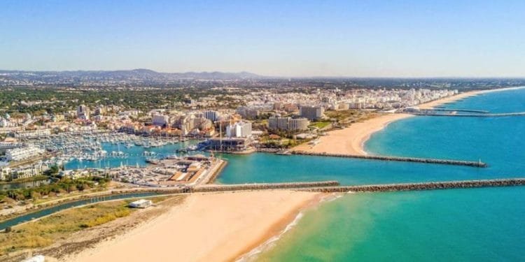 Veduta aerea di una città costiera dell'Algarve, portogallo, caratterizzata da un grande porto turistico con numerose barche ormeggiate, spiagge sabbiose che si estendono lungo la costa e un mix di edifici sparsi in tutta la città. L'oceano è di un turchese vibrante e il cielo è limpido, ideale per avventure in multiproprietà.