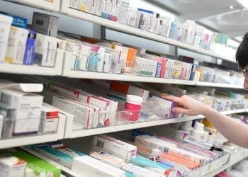 Si vede una persona che prende una scatola di farmaci da uno scaffale ben organizzato rifornito di varie forniture mediche e farmaci. Gli scaffali, che ricordano quelli portoghesi, contengono scatole colorate di varie prescrizioni o farmaci da banco in un ambiente farmaceutico.