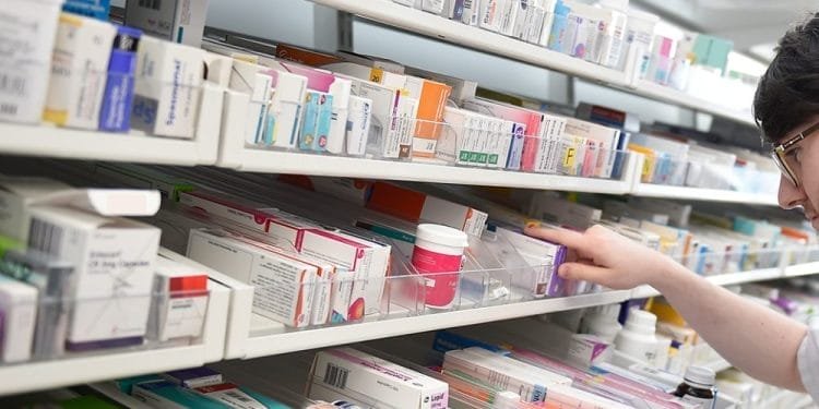 Si vede una persona che prende una scatola di farmaci da uno scaffale ben organizzato rifornito di varie forniture mediche e farmaci. Gli scaffali, che ricordano quelli portoghesi, contengono scatole colorate di varie prescrizioni o farmaci da banco in un ambiente farmaceutico.