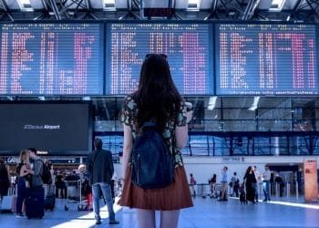Una persona con lunghi capelli scuri e uno zaino blu guarda un grande tabellone delle partenze dell'aeroporto che mostra le informazioni sul volo. Altri viaggiatori, tra cui giovani italiani all'estero, si muovono per il terminal, alcuni con bagagli. Il terminal ha soffitti alti con un orologio che segna le 12:08 sullo sfondo.