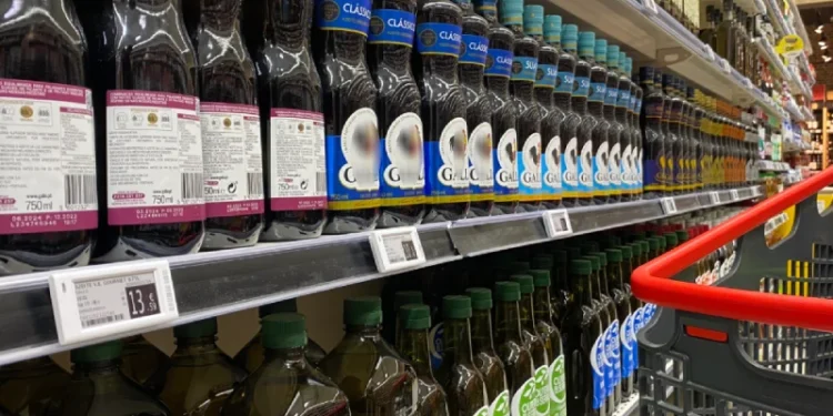 La corsia di un supermercato con file di bottiglie di olio d'oliva sugli scaffali. Le bottiglie variano per marca e colore dell'etichetta, inclusi verde, blu e viola. Un carrello rosso è parzialmente visibile sul lato destro dell'immagine. Le etichette dei prezzi (prezzo) sono visibili sotto le bottiglie, ricordando un tipico mercato portoghese (Portogallo).
