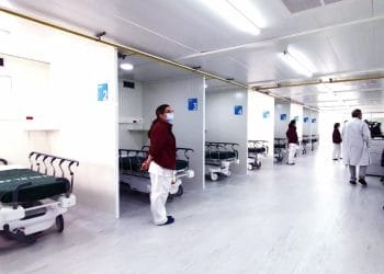 Un reparto ospedaliero con diverse aree separate, ciascuna contenente un letto ospedaliero e attrezzature mediche. Il personale medico, alcuni in camice bianco e altri in uniformi marrone, sono in piedi e camminano in giro. L'ambiente è pulito e ben illuminato, ricorda un pronto soccorso ben organizzato.