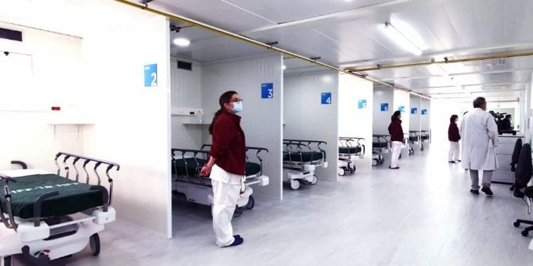 Un reparto ospedaliero con diverse aree separate, ciascuna contenente un letto ospedaliero e attrezzature mediche. Il personale medico, alcuni in camice bianco e altri in uniformi marrone, sono in piedi e camminano in giro. L'ambiente è pulito e ben illuminato, ricorda un pronto soccorso ben organizzato.