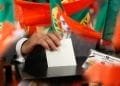 Una persona esprime un voto mettendo una scheda bianca in una scatola. Molteplici bandiere rosse e verdi, probabilmente portoghesi a causa del loro disegno, circondano la scena. La persona invece è in possesso di una carta d'identità del Portogallo e partecipa al votare elezioni Portogallo.