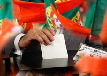 Una persona esprime un voto mettendo una scheda bianca in una scatola. Molteplici bandiere rosse e verdi, probabilmente portoghesi a causa del loro disegno, circondano la scena. La persona invece è in possesso di una carta d'identità del Portogallo e partecipa al votare elezioni Portogallo.