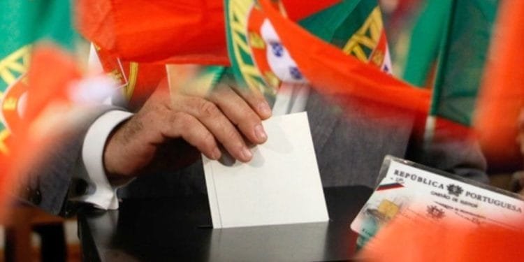 Una persona esprime un voto mettendo una scheda bianca in una scatola. Molteplici bandiere rosse e verdi, probabilmente portoghesi a causa del loro disegno, circondano la scena. La persona invece è in possesso di una carta d'identità del Portogallo e partecipa al votare elezioni Portogallo.